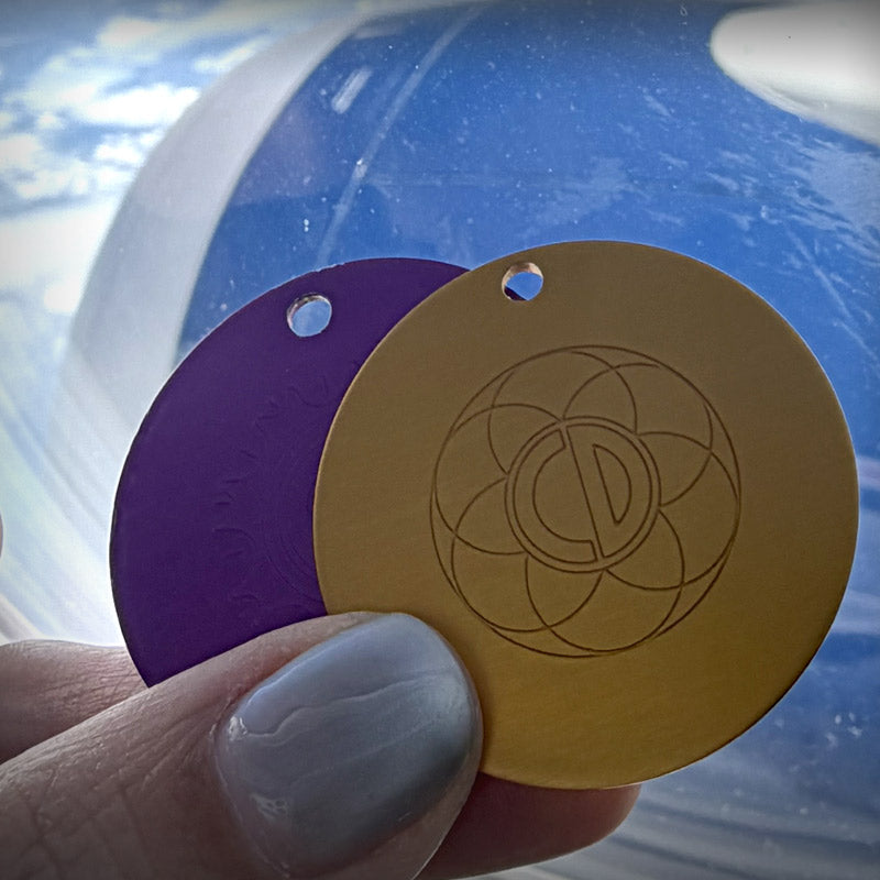Auric Illumination Disc. Purple and gold circular tags with a design held by a hand against a blue background.